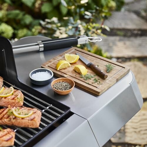 Outdoor grill with salmon, lemon slices, and herbs on a cutting board.