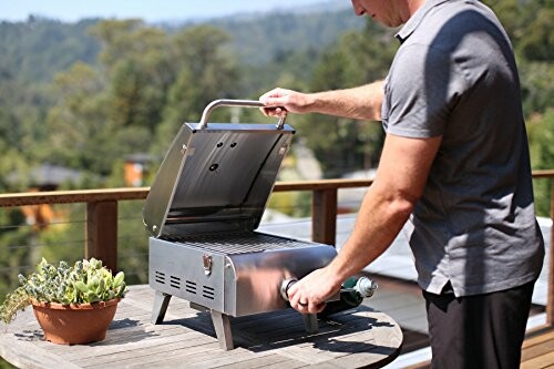 Man using a portable gas grill on a deck with a scenic background.