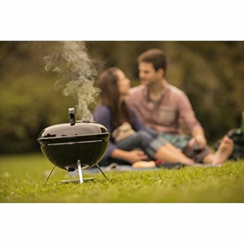 Couple sitting on grass with a smoking portable grill in the foreground.